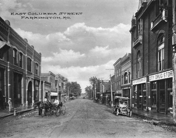 View of the East Columbia Street business district, lined with storefronts, horse-drawn carts and automobiles. Business signs read: "City Drug Store" "Hoy Grocery Co." and "Book Store." Logo on front of automobile reads: "Buick." Caption reads: "East Columbia Street Farmington, Mo."