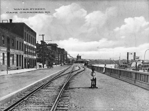 View of Water Street. Railroad tracks run along the river and split in the distance. A girl looks out at a steamboat that is visible in the river to the right. Business signs read: "Bahn Bros. Hardware" and "I. Ben. Miller Ice Cream Factory." On the right a riverboat is docked. The train depot is in the background. Caption reads: "Water Street, Cape Girardeau, Mo."