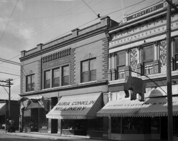 Stores in the shopping district. Business signs read: "Laura Conklin Millinery," "Dr. Wilfley Surgeon" and "Jumbo Clothing and Shoe Co." The Jumbo store has an elephant-shaped electric sign hanging over the awning. Text on buildings reads: "Young Men's Christian Association" and "Wright 1901."
