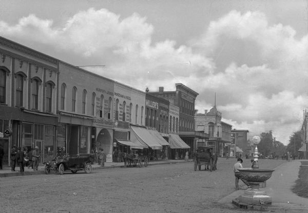 View of the West side of the town square, with automobiles, horse-drawn carts, pedestrians and a fountain. The street is lined with storefronts including "Billiards and Pool," "J.F. Ecker," "Murphy & Jones Real Estate & Farm Loans," "McKeehan & Reed," "Miller's Store," "C.A. Robinson Merc. Co.," "Willard Hotel" and "Gem."