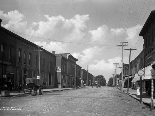 A view down Second Street, with horse-drawn carts, pedestrians and storefronts. Business signs on left read: "The Racket I. Robi," "Electric Theatre" and "Dr. Newland Dentist." Clock on right side of the street reads: "B.B. McCormick Optician."