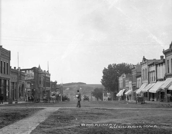 A man walks across the unpaved street in the Broadway Street business district, with automobiles and horse-drawn vehicles along the curb. There is a hill in the background. An advertisement for the circus is on a building on the left. Caption reads: "Broadway, Audubon, Iowa."