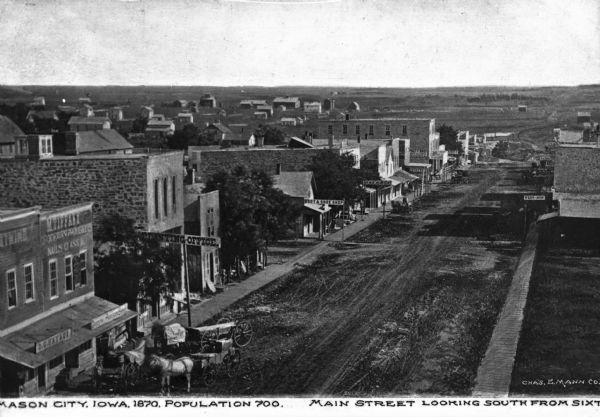 Elevated view of Main Street and shops. Business signs read: "D.C. Hay & Co.," "M. Tiffany," "Boot & Shoe Shop," "Bakery" and "West & Son." A residential area is in the distance. Caption reads: "Mason City, Iowa, 1870, Population 700. Main Street looking south from sixth."