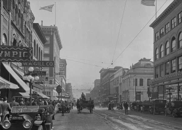 Busy street with automobiles, pedestrians and a horse-drawn cart. Streetcar tracks are in the center of the street. Business signs read: "W.A. Gill Co.," "The Rexall Store," "Will H. Beck Co. Jewelers," "Martin Fireproof Hotel," "Authier Style Shop," "Northwestern National Bank" and "Davidson Bros Co Dry Goods."