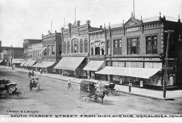 A view down South Market Street, with horse-drawn carts and a cyclist riding a "high wheel" bicycle. Business signs read: "Baldauf-Rosenblatt Co." and "Oskaloosa Savings Bank." Text on building on far right reads: "1890 Baldauf." Caption reads: "South Market Street From High Avenue, Oskaloosa, Iowa."