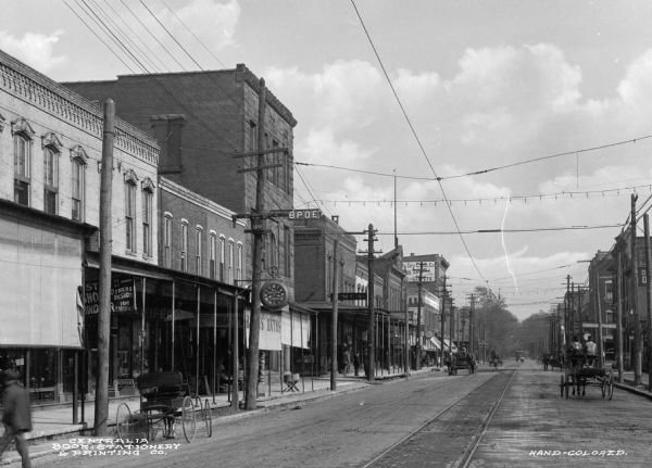 View of the East Broadway Street business district, with horse-drawn carts and cable car tracks and lines. Text on clock on left reads: "W.L. Derleth Jeweler." Sign on building on left side of street reads: "B.P.O.E."
