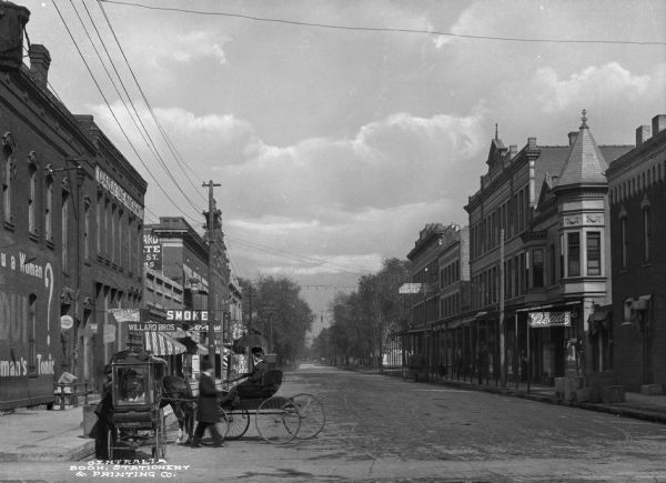 Northern view of the Locust Street business district, with a horse-drawn cart and pedestrians in the foreground, and storefronts on the left and right. Business signs read: "Electric Light & Power," "Steads," "Daily Democrat," "Willard Bros.," "Smoke," "The Club Billiards And Pool," "Plumbing," "Singer Sewing Machines" and "Special Today Only Oliver Twist."