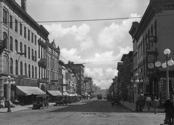 View of the Stevenson Street business district. Automobiles and horse-drawn carts line both sides of the street. Business signs read: "Dr. R.J. Burns Physician & Surgeon," "Hoss & Irving," "Dr. Torey," "Dr J.J. Grant Physician & Surgeon" and "State Bank."