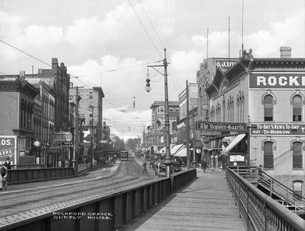 The West State Street shopping district. Electric lights and power lines are overhead. Business signs read: "The Register-Gazette," "Edison Light" and "Wearing Apparel CREDIT Man, Woman and Child."