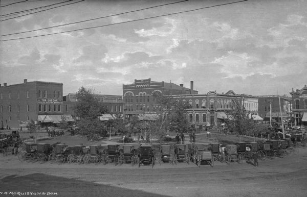 View of the northeast corner of the square. Horse-drawn vehicles are parked along the street. Business sign on left side reads: "Tom Gordon Harness, Buggies, Blankets."