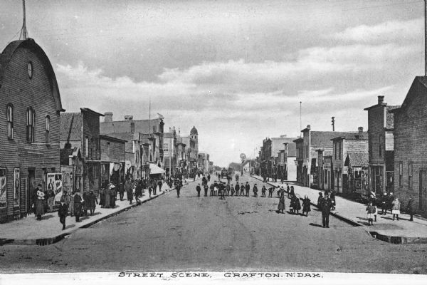 A street scene. Pedestrians are on the sidewalks and lined up in the middle of the street. Caption reads: "Street Scene, Grafton, N. Dak."