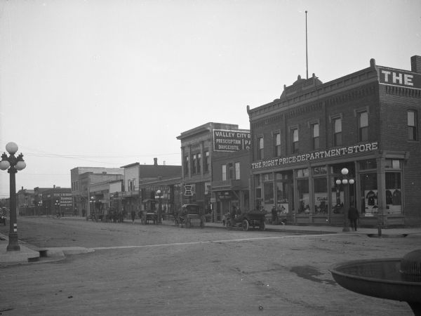 View of a shopping district. Business signs read: "The Right Price Department Store," "Valley City (obscured) Prescription Druggists," "Meat Market," "The Fair," "Hub Pool Room," "Short Order Restaurant" "Wardrobe" and "Berg & Benson."