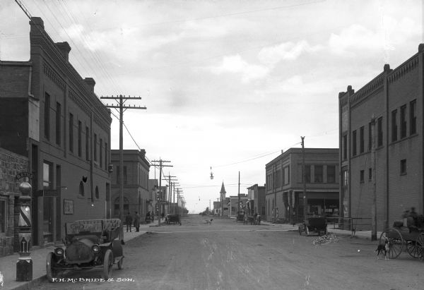 A view of West Second Street. Automobiles and a horse-drawn vehicle are parked on the street. A barber's pole reading: "Baths" is on the left side of the street. Other business signs read: "H.B. Senn Advocat" and "Bakery Lunch Room."
