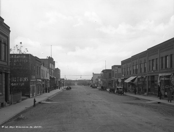 View of Main Street. Business signs read: "Rugby Confectioner," "The "Grill" Cafe," "Lyric," "Drugs," "Millinery" and "Mrs. Fredricksen Naturopath."