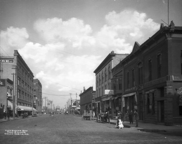 View of Demers Avenue. A woman and child and other pedestrians are on the street. Business signs read: "E.J. Lander & Co,." "Johnson & Nisbet Groceries," "Logan's Cafe" and "R.B. Griffith."