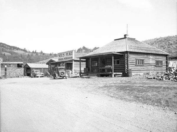 A view of a village street and general store. Business sign reads: "Neil's Place General Merchandise." A gas pump and metal sign are on the left.