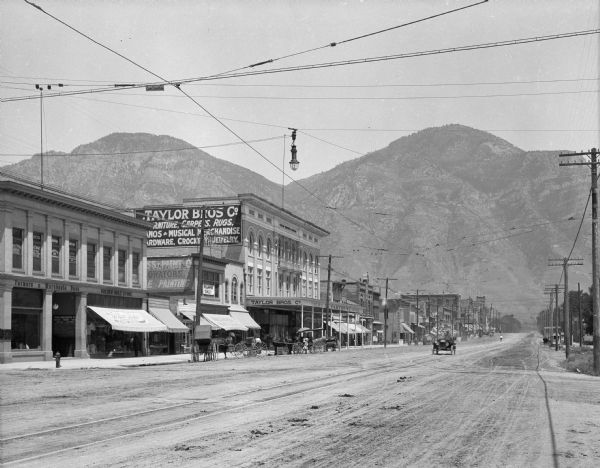 View of a street lined with storefronts and utility lines. An automobile is being driven down the street. Business signs read: "Farmers & Merchants Bank," "Golden Rule Store" and "Taylor Bros. Co." Mountains are in the background.