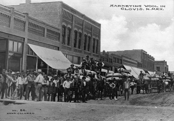 A street scene featuring a large group of men gathered around wagons loaded with bags of wool for marketing. Business signs read: "Mandell Quinn & Co.," "Kendall Dry Goods Co." and "The Hub." Caption reads: "Marketing Wool In Clovis, N. Mex."