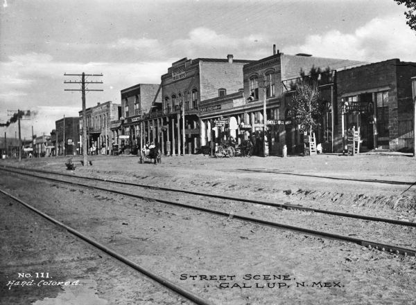 Smoke from a train is on the left side. Business signs read: "New York Store Dry Goods," "O'Neill's Shoe Shop," "Post Office," "CDI Son & (obscured) Phonograph records," "Palmer-Ketner Gen. Merchandise," "Kitchen's Opera House," "Restaurant" and "Pool." Caption reads: "Street Scene. Gallup, N. Mex."