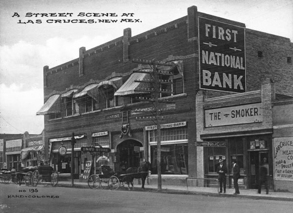 Pedestrians and horse-drawn vehicles are on the street in front of several storefronts. Business signs read: "Las Cruces Meat Co. Eggs & Poultry Fish & Game In Season," "The Smoker," "Billiards Pool And Cigars," "First National Bank," "Dentist" and "Drug Store." Caption reads: "A Street Scene At Las Cruces, New Mex."