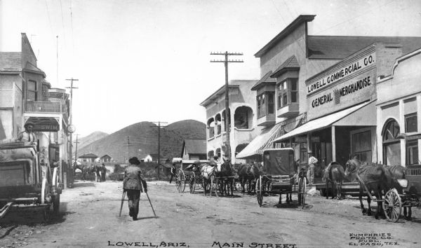 A man on crutches is walking down the street. Horse-drawn vehicles are parked along the street. Business sign reads: "Lowell Commercial Co. General Merchandise." Housing and mountainous countryside are in the distance. Caption reads: "Lowell, Ariz. Main Street."