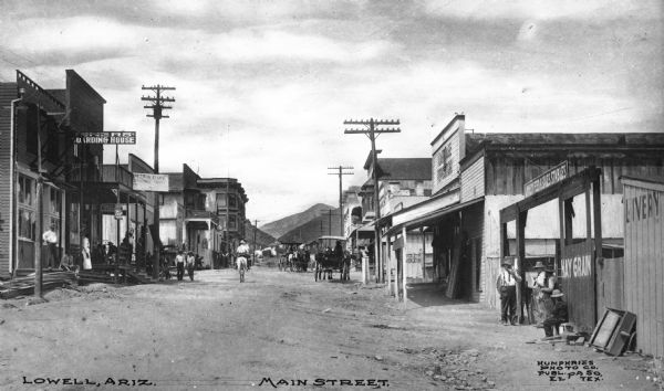 View of Main Street. Pedestrians, horse-drawn vehicles and a man on horseback are in the street. Business signs read: "(obscured) Boarding House," "Pete's Cafe" and "Livery Feed & Sales Stables." Caption reads: "Lowell, Ariz. Main Street."