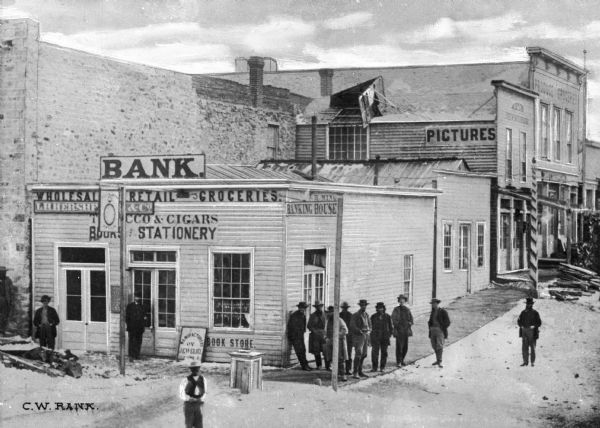 Virginia City was the first capitol of Montana. Elevated view of a group of men standing near a building with a sign reading: "L.H. Hershe (obscured) & Co.," which appears to be a bank and general store. Another business sign reads: "Pictures." A barber's pole is nearby.