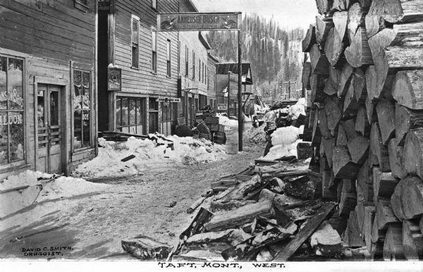 Street scene during winter. The town is no longer in existence; it sprang up during construction of the Chicago, Milwaukee & St. Paul Railroad and had the reputation of being the "toughest town in the U.S." The town, named for President Taft, was located between Saltese, Montana and Mullan, Idaho. A wood pile is near the storefronts, which appear to be taverns. Business signs read: "Swanson Bros. Saloon" and "Anheuser-Busch St. Louis Beer." Caption reads: and "Taft, Mont., West."
