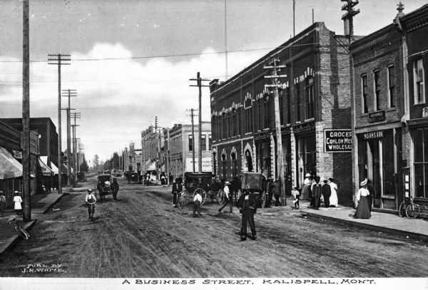 Horse-drawn vehicles are parked along the roadside and pedestrians are walking and riding bicycles on the unpaved street. Business signs read: "Noah's Ark" and "Conlon Mercantile Co." Caption reads: "A Business Street, Kalispell, Mont."