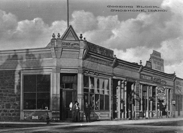 Gooding Block shopping area. Two men are standing in front of a store. Business signs read: "Stock Growers Mercantile Co.," "General Merchandise," "First National Bank," "T.M. Starrh," "Carey Lands" "Land Office Idaho Irrigation Co. Fruit and Farm Lands," "Drugs Soda" and "Burkill's" (rest of sign obscured). Caption reads: "Gooding Block, Shoshone, Idaho."