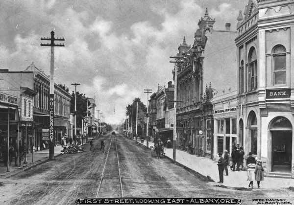 View of the First Street business district looking east. Pedestrians and horse-drawn vehicles are in the street. Business signs read: "Bank" "Drug Store," "A.M. Freerksen Justice of the Peace," "Western Union Telegraph Office," "Wells Fargo & Co. Express," "Overland Ticket Office," "Books Stationery," "Circulating Library" "and Grocery"(obscured) and "Barber Shop" (obscured). Caption reads: "First Street, Looking East-Albany, Ore."
