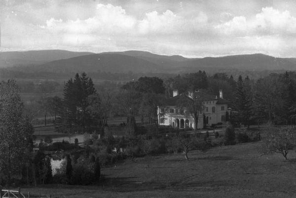 View of a large residence surrounded by trees. The mansion sits on a large piece of property with a pond, and a mountainous countryside is in the distance.