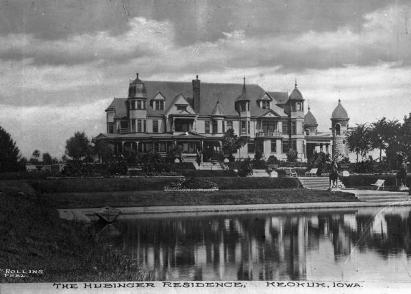 View across pond toward the J.C. Hubinger mansion. Two stag statues stand by the pond. J.C. Hubinger, a prominent Iowa businessman, opened a starch factory in Keokuk in 1887. Caption (partially obscured) reads: "The Hubinger Residence, Keokuk, Iowa."