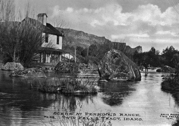 View of Perrine's Ranch, located in the vicinity of Twin Falls. A man is crossing a stream filled with large rock boulders. The ranch is on the left side of the stream and mountains are in the background. Caption reads: "Scene At Perrine's Ranch Twin Falls Tract, Idaho."