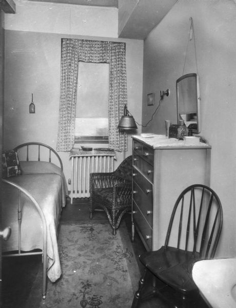 View of a bedroom in Tatham House YWCA. Small room with a single bed, sink, dresser, chairs and a lamp.