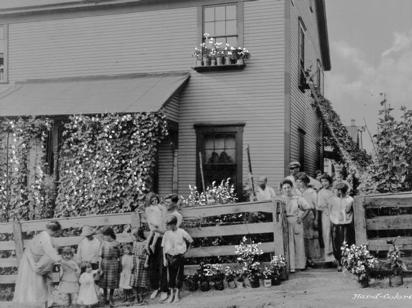 A large group of adults and children pose near a home in a company town of Consolidated Coal Company. On the right side of the home vines are growing on a large trellis.