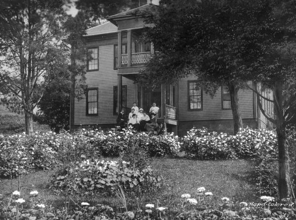 A group of eight women poses on the porch of a two-story home. The yard in front of the home is filled with flowers. Taken in a company town of Consolidated Coal Company.