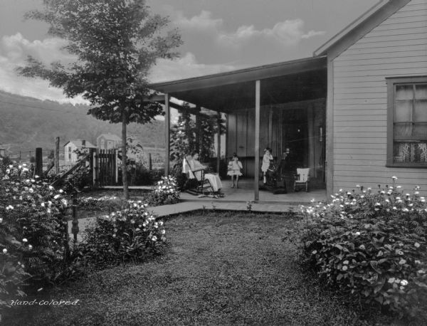 A woman and two children sit in the porch area of their home.  One of the children plays on a rocking horse.  A groomed yard and flowering shrubs are in front of the porch. A neighboring homestead is in the background. Photograph taken in company town of Consolidated Coal Company.