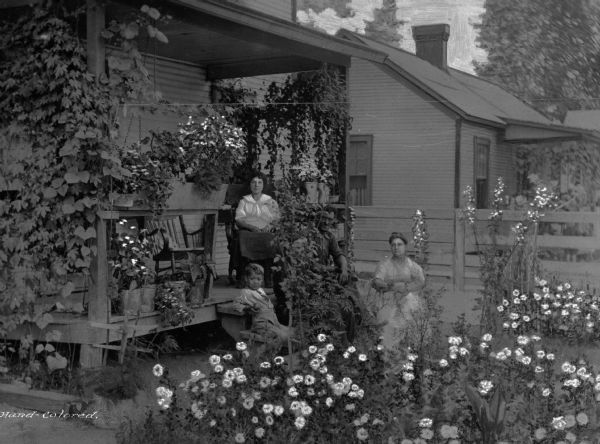 A man, two women and a little boy sit in the porch and garden area of a home. Fairmont was a company town of Consolidated Coal Company. Flowering plants are in the yard, and there are many plants in the porch area. A neighboring home is next door.