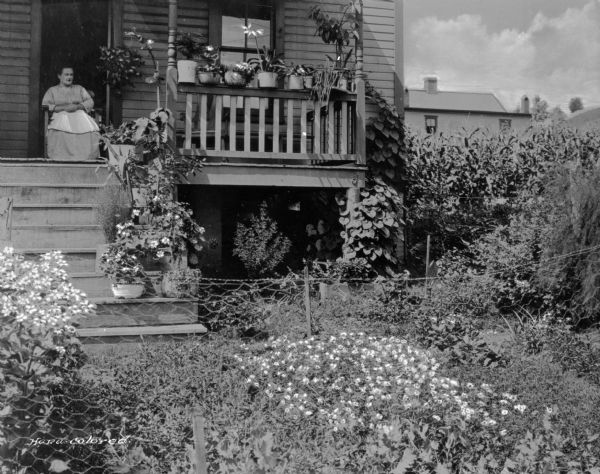 A woman sits on the back porch of a home in a company town of Consolidated Coal Company. There are many plants on the porch, and a garden in the yard. A cornfield and neighboring home is behind the house.