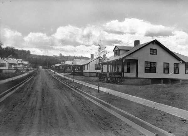View of a street with largely identical newly built houses in a company town of Pepperell Manufacturing Company. The street and yards are dirt, and trees have been newly planted in front of the homes.