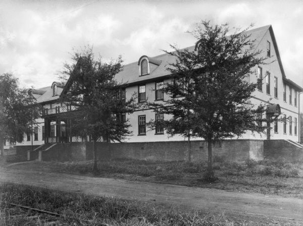 A view of a large two-story boarding house in a company town of Pepperell Manufacturing Company. There are several large trees in front of the building, which lies on a dirt road. Railroad tracks are in the left foreground.