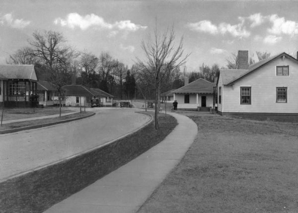A view of winding residential street lined with homes. Several cars are parked in the distance, and pedestrians are on the street. Taken in a company town of Pepperell Manufacturing Company.