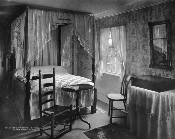 Antiquarian house bedroom interior. Room features a canopy bed with a white lace canopy, two chairs, one a Shaker style chair, two small tables, and a mirror. Caption reads: "© Antiquarian Society Plymouth, Mass."
