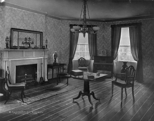 View of a room in the antiquarian house. Formal room with two windows, fireplace, mantle, mirror, chairs, tables, desk and chandelier. Caption reads: "© Antiquarian Society Plymouth, Mass."