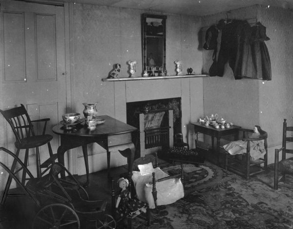 View of antiquarian house nursery. Room features a small fireplace, children's toys, dolls, clothing, furniture, a tea set, a high chair and a small wooden toy carriage.