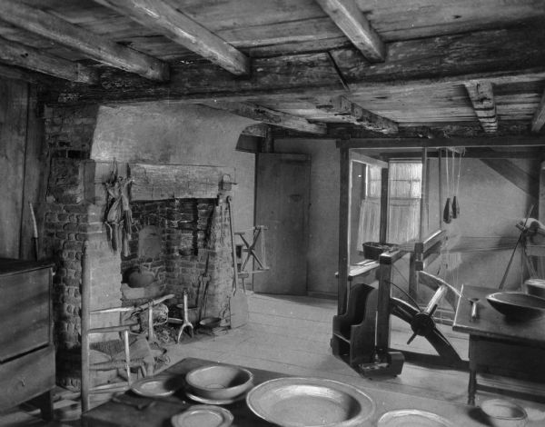View of interior of Harlow House. Built in 1677, the building was originally the residence of William Harlow and it still stands and remains open for tours and educational programs. The room features a ceiling made of wooden beams, a brick fireplace, weaving loom, dining table, tools, Shaker style chair, small table, and child's chair and toy cradle. A small swift tool is obscured by the fireplace.