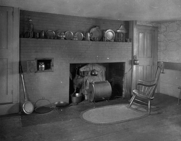 View of antiquarian house kitchen. Kitchen interior features a rocking chair, rug, large brick fireplace with cooking tools, pots and a bed warmer. There is a small smoker in the fireplace. Tableware, kettles, pots and dried herbs are on the mantle.