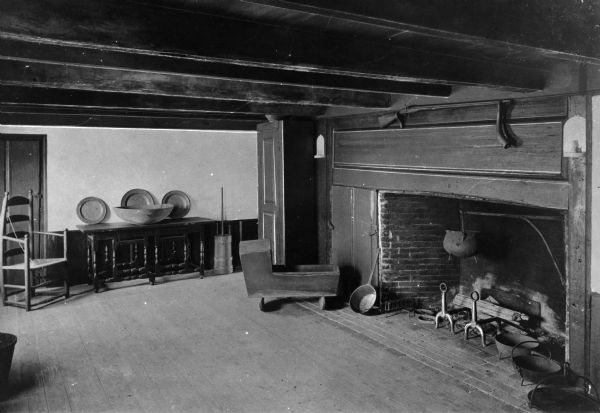 Interior view of the Major John Bradford house. Major John Bradford was the grandson of pilgrim governor William Bradford. Built in 1714, the home still stands. Room features a fireplace, cooking utensils, butter churn, Shaker style chair, a table, and a wooden cradle. A rifle is over the fireplace.