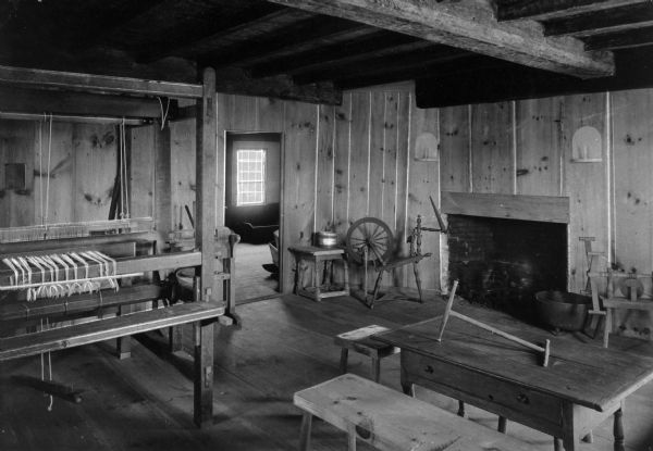 An interior view of Major John Bradford house that was built in 1714 and is still standing. Major John Bradford was the grandson of pilgrim governor William Bradford. The room features a fireplace, large cooking pot, spinning wheel, loom, yarn swift, table and benches.